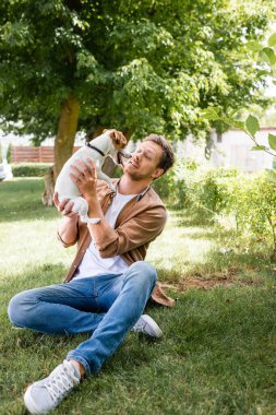 Genç adam yeşil çimenlerde otururken Jack Russell Terrier köpeğini tutuyor.