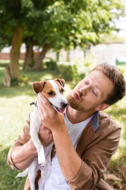 young man making duck face while holding jack russell terrier dog in park clipart
