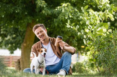 Kahverengi gömlekli ve kotlu genç adam, Jack Russell Terrier 'ın yanındaki çimlerde otururken tek kullanımlık bardakları tutuyor.