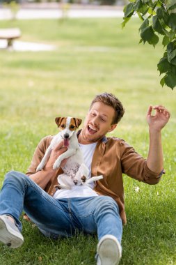Çimenlikte eğlenirken Jack Russell Terrier köpeğini tutan sıradan giyinmiş heyecanlı bir adam.