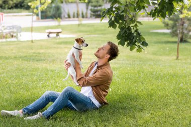 Kotlu ve kahverengi gömlekli genç adam yeşil çimlerde oturuyor ve Jack Russell Terrier köpeğini tutuyor.