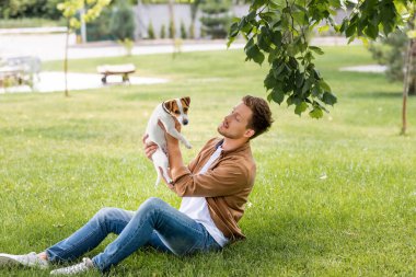 young man in brown shirt and jeans holding jack russell terrier dog while sitting on lawn clipart