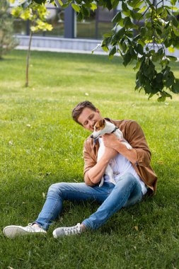 Kahverengi gömlekli ve kot pantolonlu genç adam çimenlerde oturup Jack Russell Terrier 'a sarılıyor.