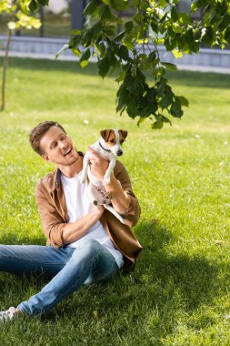 Kotlu genç adam yeşil çimlerde otururken Jack Russell Terrier köpeğini tutuyor.