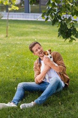 Yeşil çimenlerde oturan ve Jack Russell Terrier 'a sarılan sıradan elbiseli genç adam.
