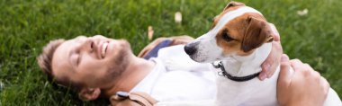 Yatay konsept, çimenlikte gözleri kapalı yatan ve Jack Russell Terrier köpeğine sarılan adam.
