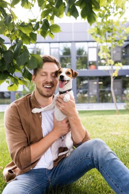 Genç adam ağaç dalının altında çimenlerde oturuyor ve Jack Russell Terrier köpeğini tutuyor.