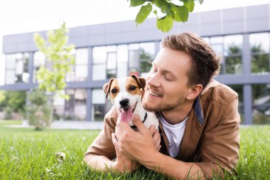 Genç adam yeşil çimlerde yatarken Jack Russell Terrier köpeğine sarılıyor. 