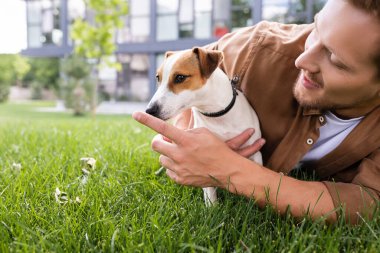 Genç adam yeşil çimlerde yatıyor ve Jack Russell Terrier 'ın burnuna dokunuyor.