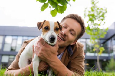 Yeşil çimenlerin üzerinde uzanmış Jack Russell Terrier köpeğine sarılan genç adamın seçici odağı