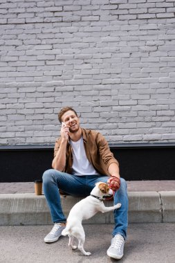 Jack Russell Terrier 'ı tasmalı tutan adam, şehir sokaklarında kahve yakınında akıllı telefonla konuşurken... 