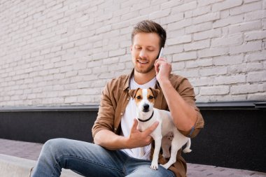 Jack Russell Terrier 'a sarılan ve binanın ön cephesinde kaldırımda konuşan adam. 