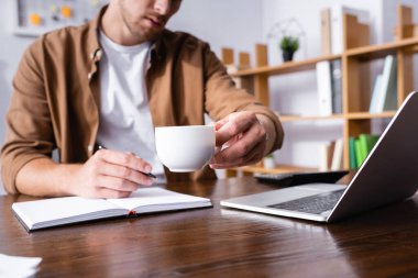 Selective focus of businessman holding cup of coffee while working with notebook near laptop  clipart