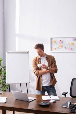 Ofiste elinde bir fincan kahve tutarken dizüstü bilgisayara bakan düşünceli iş adamının seçici odağı 