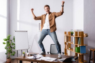 Businessman showing yeah gesture while standing near gadgets and papers on office table  clipart