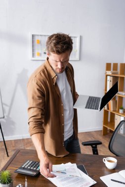 Dizüstü bilgisayar tutan işadamlarının seçici odak noktası ve ofiste çizelgeleri olan kağıtlara bakmak. 