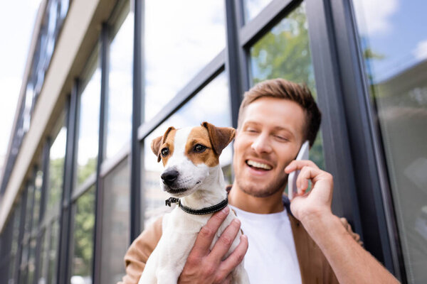 selective focus of young man talking on smartphone while holding jack russell terrier dog on street