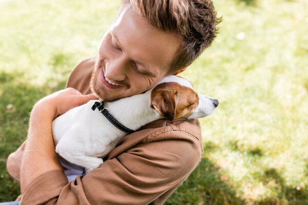 high angle view of young man with closed eyes holding white jack russell terrier dog with brown spots on head