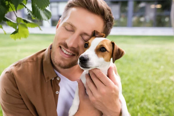 Gözleri kapalı genç bir adam beyaz Jack Russell Terrier köpeği tutuyor kafasında kahverengi lekeler var