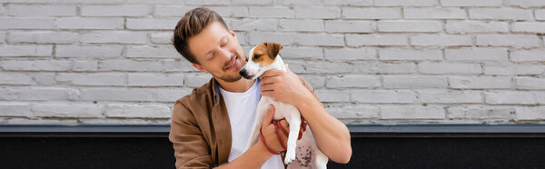 Panoramic shot of young man hugging jack russell terrier on urban street 