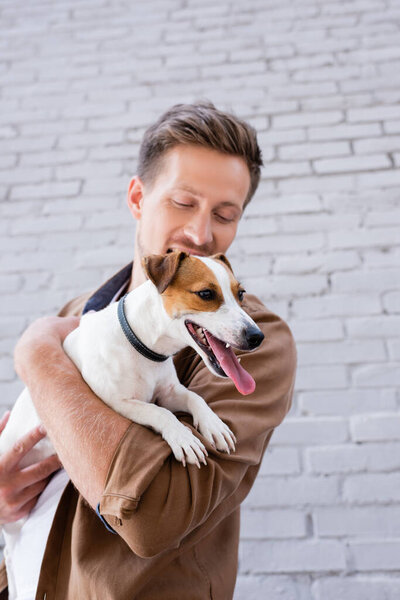 Selective focus of man holding jack russell terrier with sticking out tongue near building 