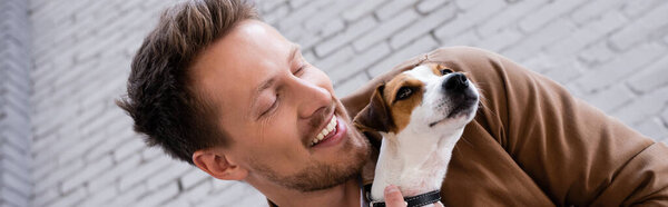 Horizontal image of man holding jack russell terrier near facade of building 