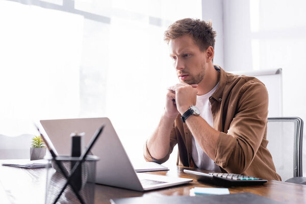 Selective focus of young businessman looking at laptop near calculator and stationery in office 