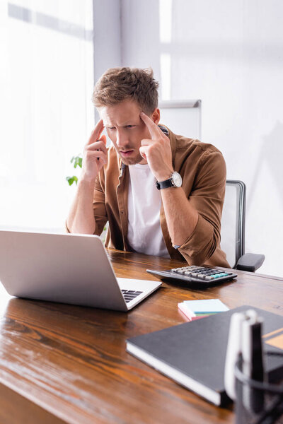 Selective focus of focused businessman looking at laptop near stationery and calculator in office 