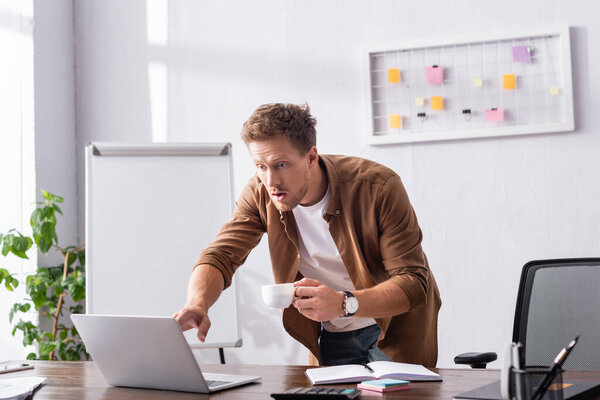 Selective focus of excited businessman with cup of coffee looking at laptop in office 