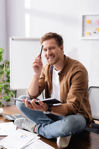 Young businessman with pen and notebook looking at camera while sitting on table in office