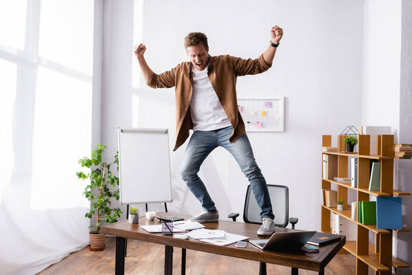 Young businessman showing yes gesture while standing on table in office 