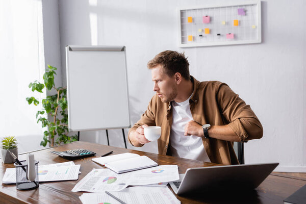 Selective focus of young businessman coughing and holding coffee cup at working table 