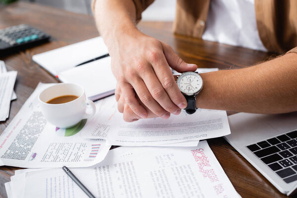 Cropped view of young businessman touching wristwatch near papers, laptop and cup of coffee on table 