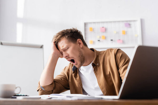 Selective focus of young businessman yawning while working in office 