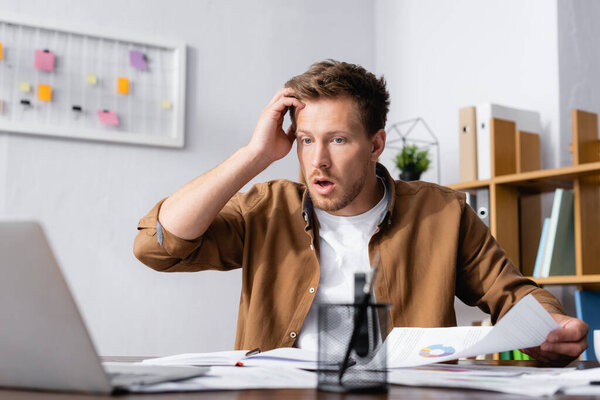 Селективный фокус шокированного бизнесмена, держащего документ и смотрящего на ноутбук в офисе 