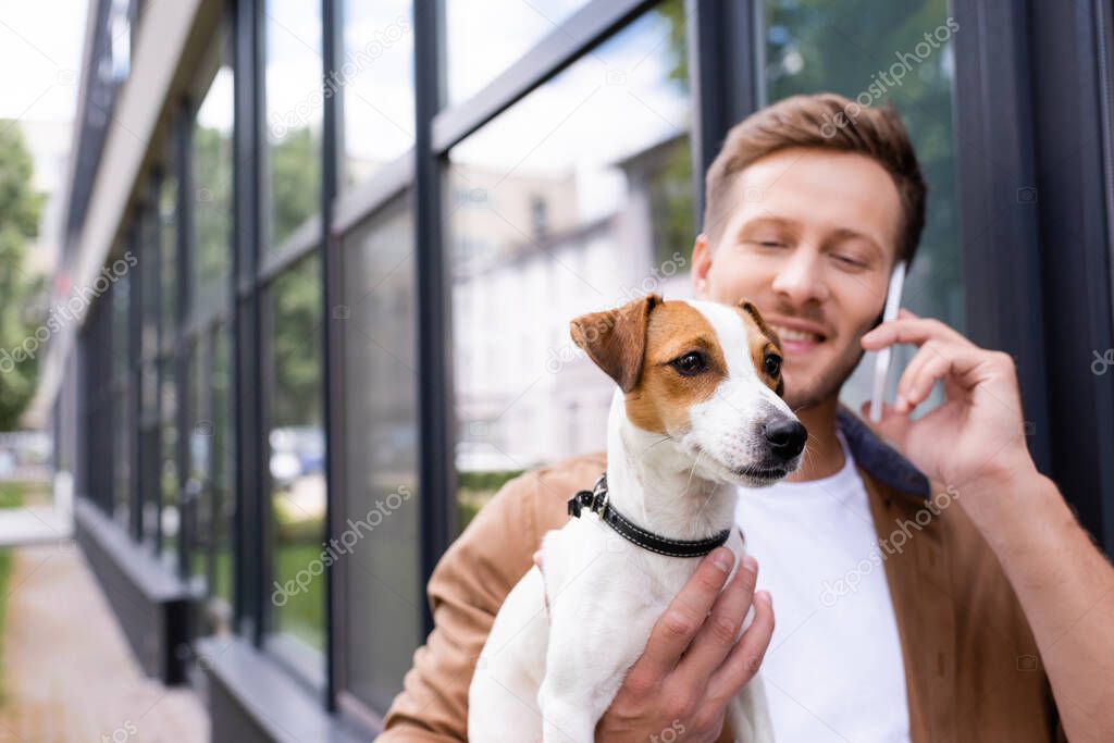 selective focus of young man holding jack russell terrier dog and talking on smartphone outdoors