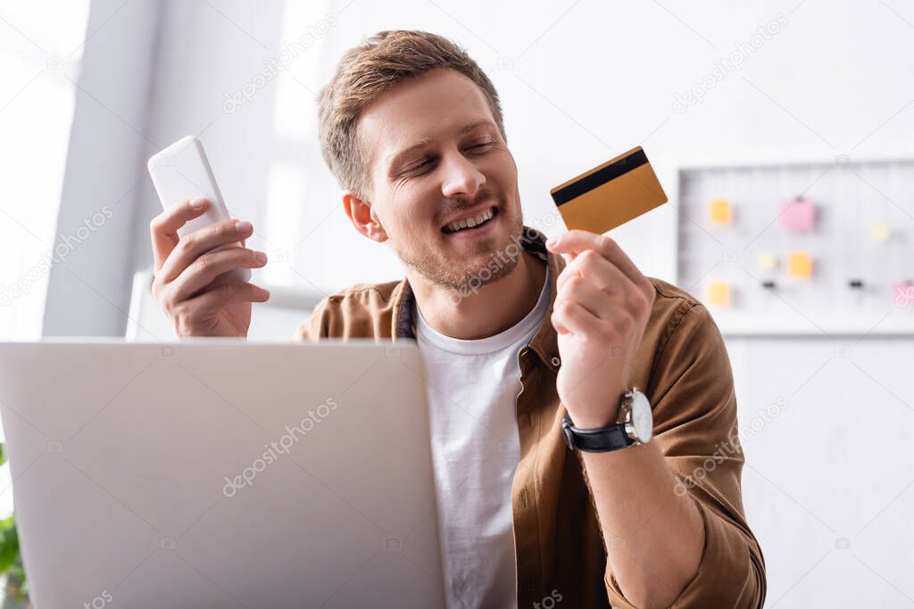 Selective focus of businessman holding credit card and smartphone near laptop in office 