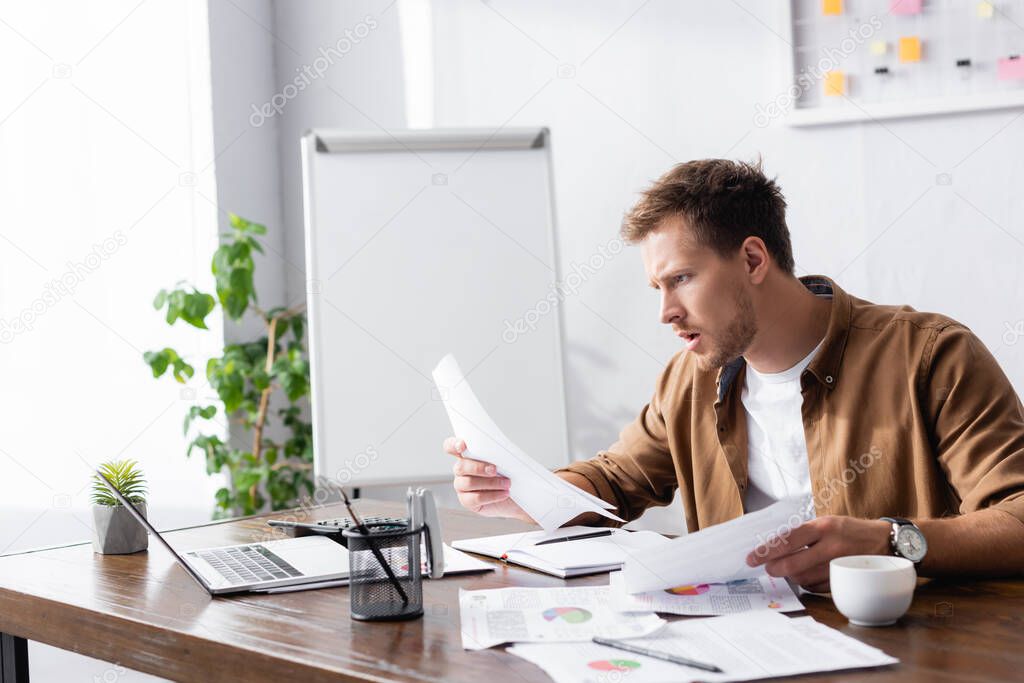 Selective focus of focused businessman working with documents near laptop and coffee on office table 