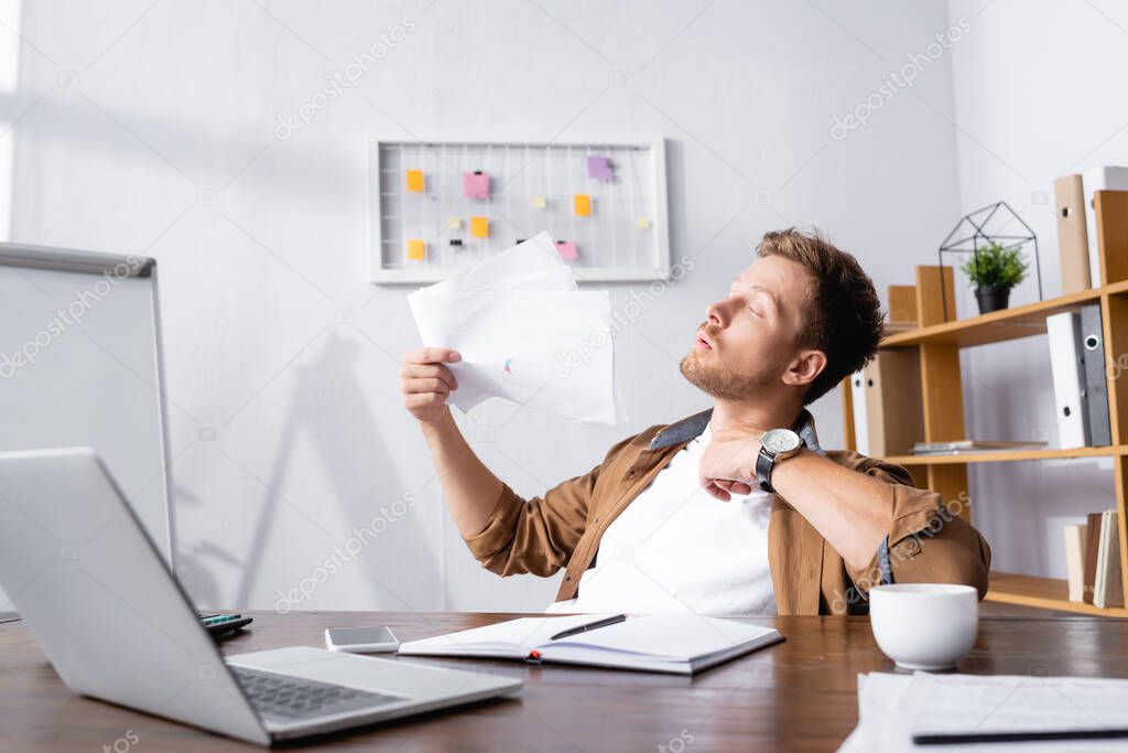 Selective focus of young businessman blowing with documents while suffering from heat at table in office 