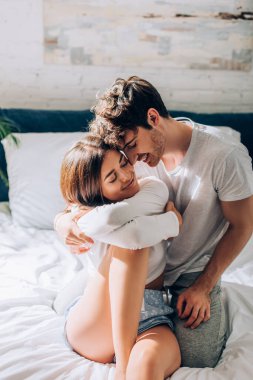 Pijamalı genç adam yatakta oturuyor ve gözleri kapalı kız arkadaşına sarılıyor.