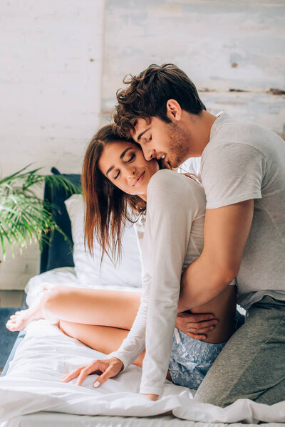 Young man with closed eyes sitting on bed and embracing girlfriend