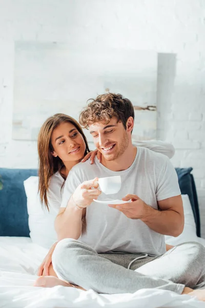 stock image Young woman sitting on bed and looking at boyfriend with cup of coffee