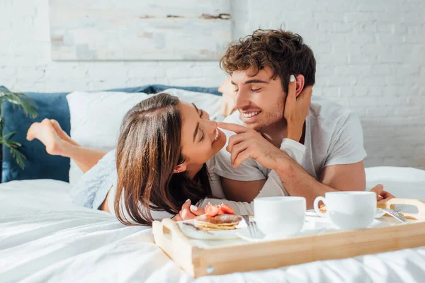 年轻夫妇在咖啡和煎饼放在床上的早餐盘旁边互相触摸的选择性焦点 — 图库照片
