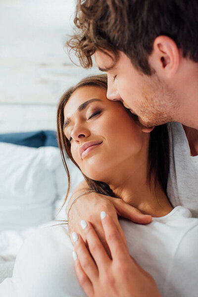 Selective focus of man with closed eyes kissing girlfriend on bed 