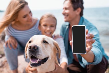 Seçici odak noktası, aile ve Golden Retriever plajlarında boş ekranlı akıllı telefon tutan adam. 