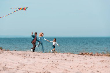 Ailenin seçici odak noktası deniz kenarında uçurtmayla koşarken el ele tutuşmak. 