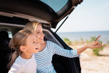 Seçici odaklı bir kız, annesinin yanından uzaklaşıp kumsaldaki parmak ve arabayı işaret ediyor. 