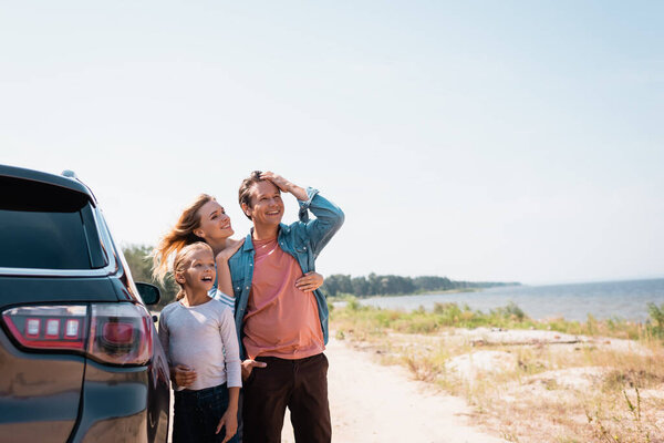 Селективное внимание женщины, обнимающей мужа и взволнованную дочь возле машины на берегу моря 