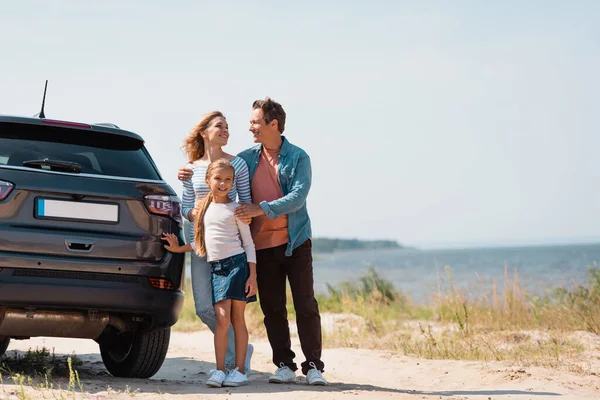 Babanın, kızını karısına ve arabasına sarılışının seçici odak noktası deniz kıyısında bir plaj. 