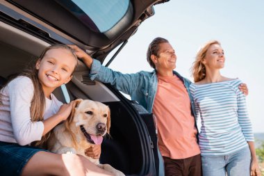 Golden Retriever 'ı okşayan kızın seçici odak noktası açık havada arabanın yanında kucaklaşan ebeveynlerin yanı. 